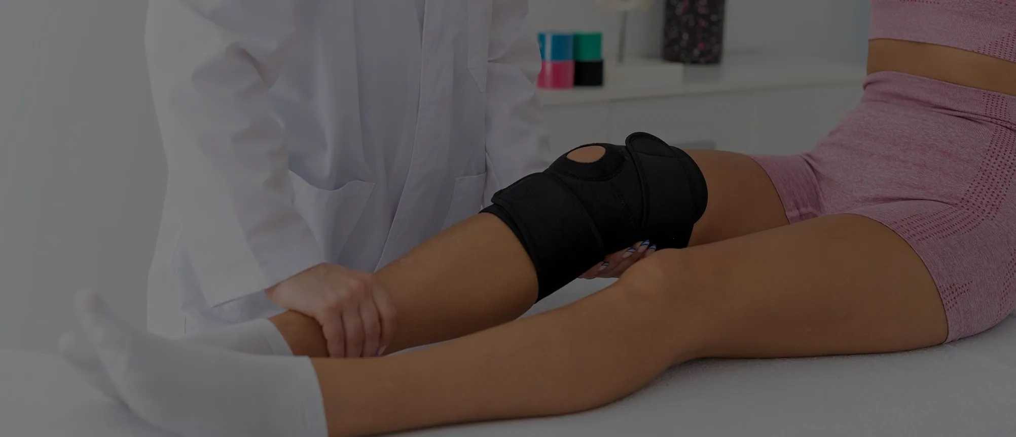 Une personne en tenue médicale examine le genou d'une patiente portant une genouillère noire et un ensemble de sport rose.