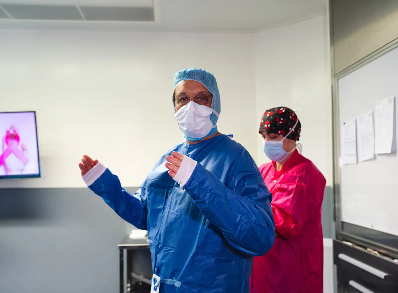 Deux professionnels de santé en salle blanche, portant des masques, dont un en tenue bleue de chirurgie avec charlotte et lunettes.