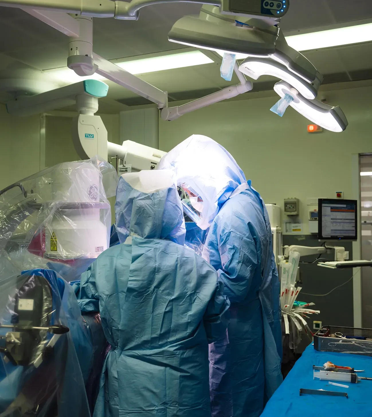 Deux chirurgiens en tenue stérile bleue et protections faciales effectuent une intervention chirurgicale sous une lampe opératoire dans une salle d'opération équipée de matériel médical.